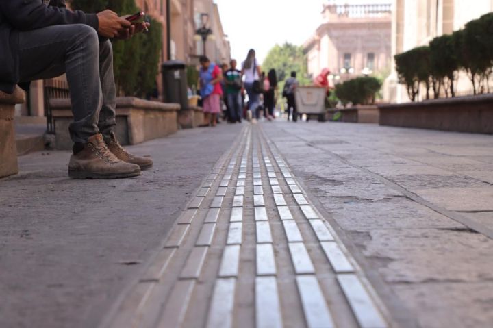 Imagen de una guía podotáctil en una calle del Centro Histórico de San Luis Potosí, con una persona sentada en un banco y varios peatones al fondo.