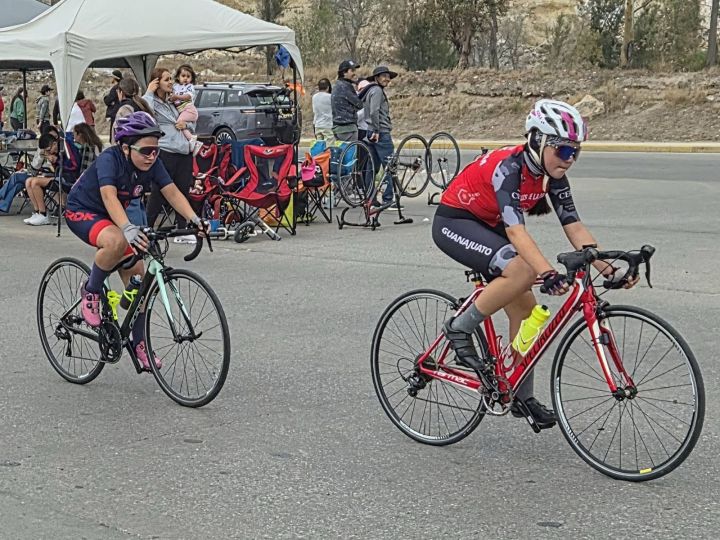 Dos jóvenes ciclistas compiten en una carrera, una en bicicleta verde y la otra en bicicleta roja. Al fondo, hay espectadores y una carpa.